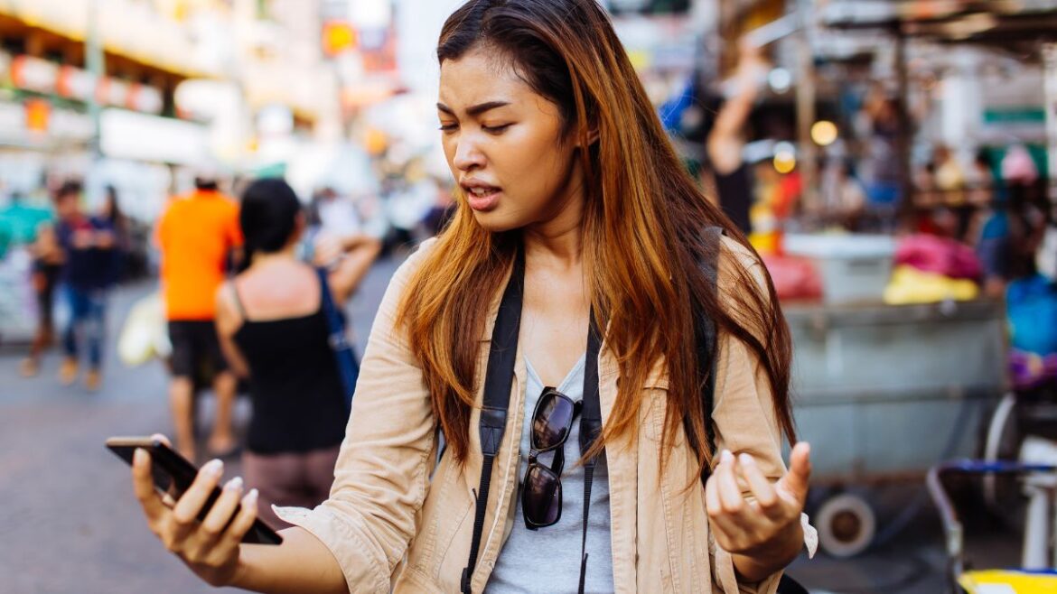 Young female traveler looking at phone outside on street with displeased face