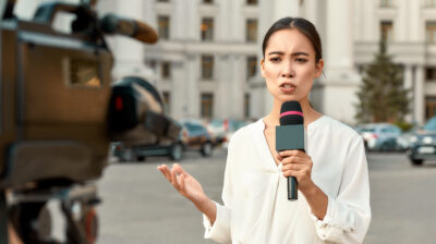 Cropped portrait of professional female reporter at work. Young woman standing on the street with a microphone in hand and smiling at camera. Horizontal shot. Selective focus on woman